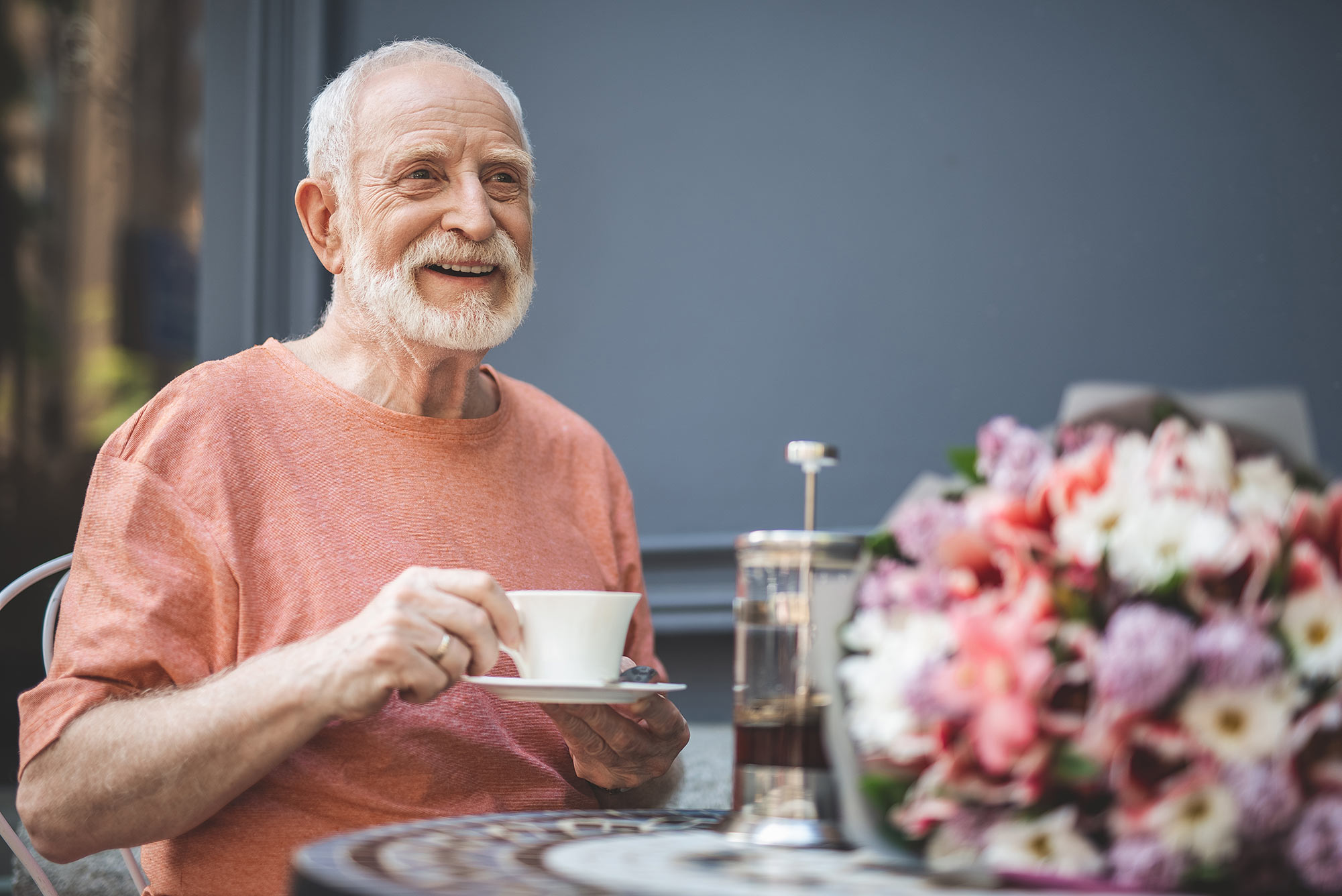Ein lächelnder älterer Mann sitzt an einem Tisch auf dem ein Blumenstrauß steht und trinkt Tee